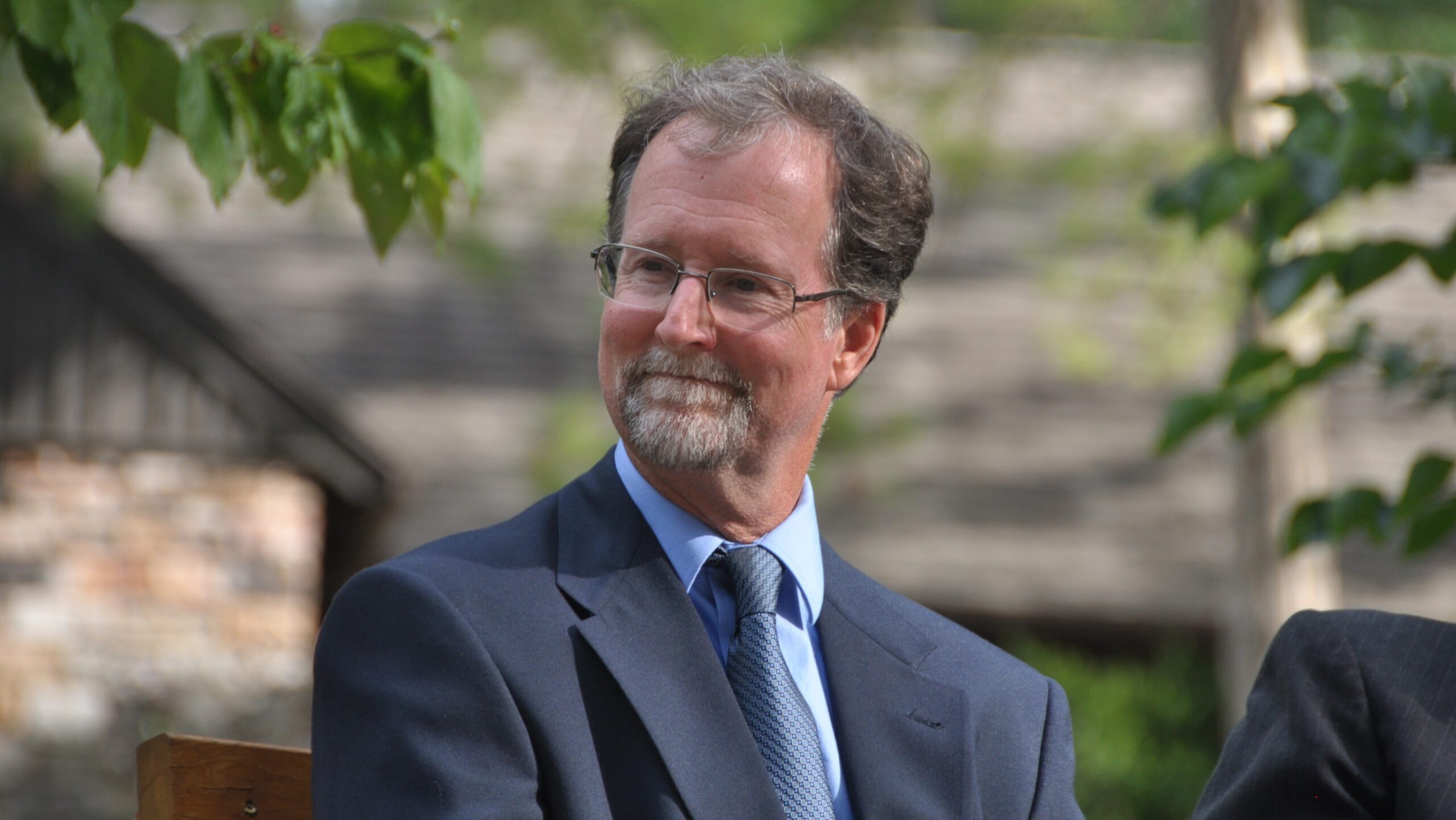 A man in a suit looks to his right while standing among green leaves and a wooded backdrop.
