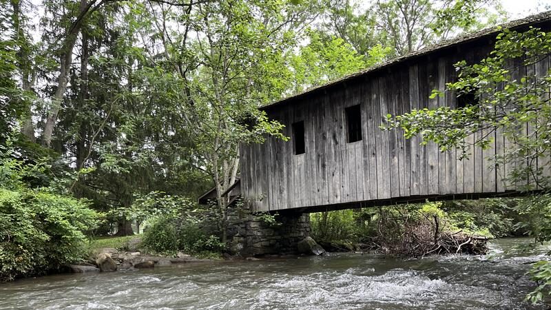 A covered wooden bridge crosses a stream in a green forest.