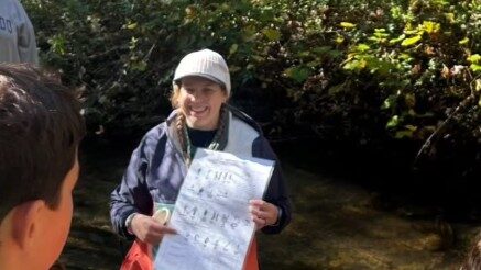 Haley standing in a stream holding a poster and giving instructions to a group of people.
