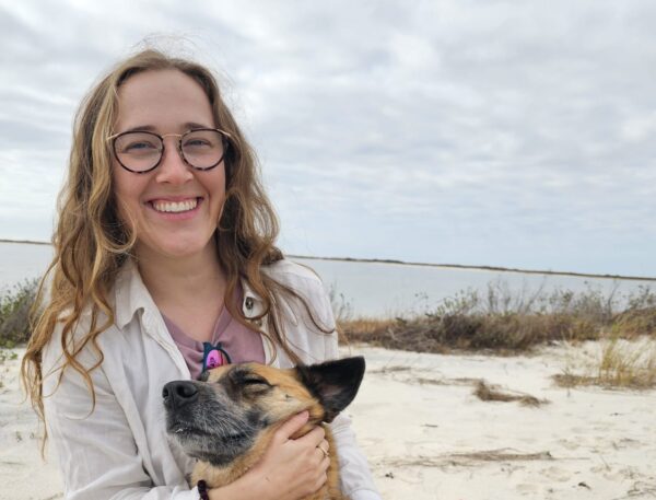 Rory hugs her dog while sitting on a beach with dunes in the background.