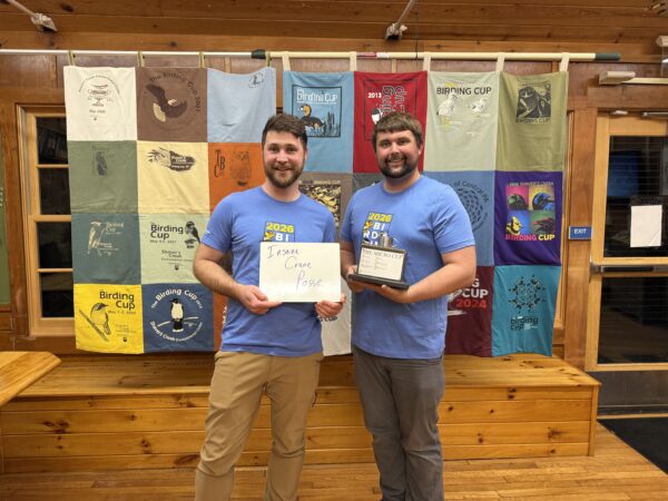 Two birders stand in front of quilted wall hanging holding a bird watching award