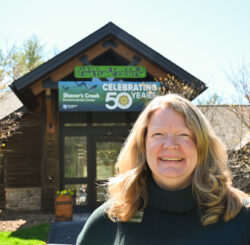 A person with shoulder-length blonde hair stand smiling in front of a wooden building with a blue sky and trees in the background