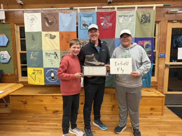 Three birders stand in front of quilted wall hanging holding a bird watching award