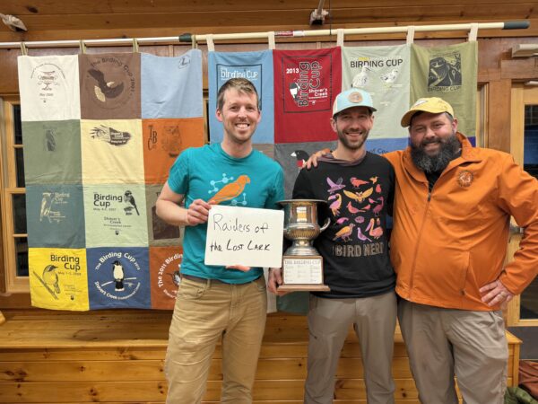 Three birders stand in front of quilted wall hanging holding a bird watching award