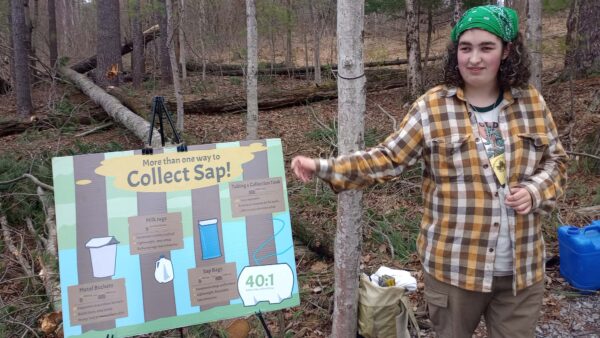 Catherine, wearing a bandana on her head and a flannel shirt, gestures toward a poster about the maple sugaring process during the Shaver's Creek Maple Harvest Festival.