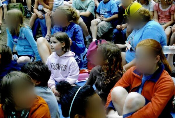 A young girl wearing a pink hooded sweatshirt sits with other children at a summer camp.