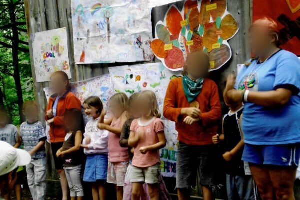 A young girl with a pink hooded sweatshirt stands with her camp friends in front of drawings made my campers.