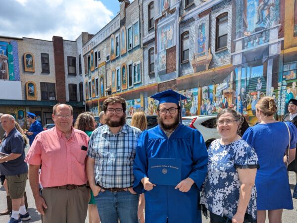 A family of four poses in front of a building with artwork on the front. One of them is wearing a graduation cap and gown and holding a diploma.