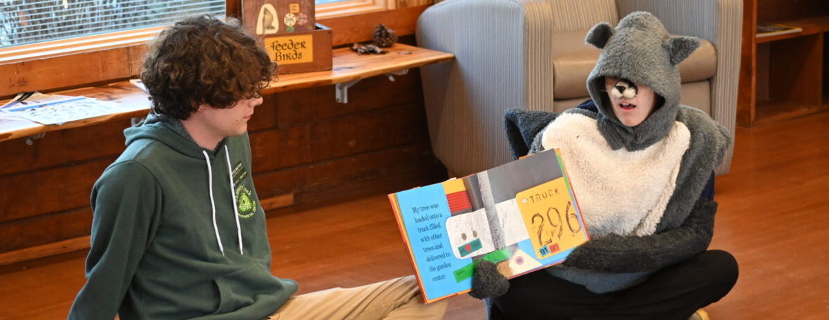 Kids Corner participants sit on the floor and watch a person dressed in an animal costume read a picture book.