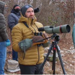 Andrew Bechdel wearing a beanie and a puffy jacket and standing in the forest with a spotting scope.