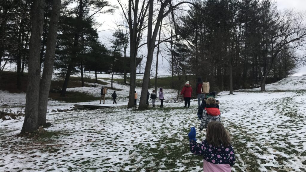 An adult leads a group of children across a snowy patch of grass toward some trees.