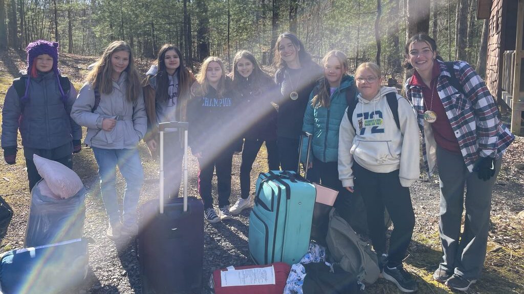 A group of Outdoor School students stands near their luggage on a gravel path leading through the forest.