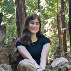 A girl in a brown shirt sits on the ground among trees.