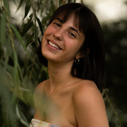 A woman with dark hair smiles at the camera near green leaves.