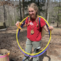 A girl in a red shirt smiles at the camera while holding a yellow hula hoop around her neck.