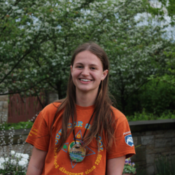 A girl in an orange shirt smiles at the camera with trees in the background.