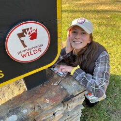 A woman in a hat crouches next to a sign that says "Pennsylvania Wilds"