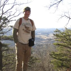 A man in a white shirt stands among the trees with an overlook in the background.