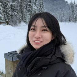 A girl in a black jacket smiles at the camera with a snowy scene in the background.