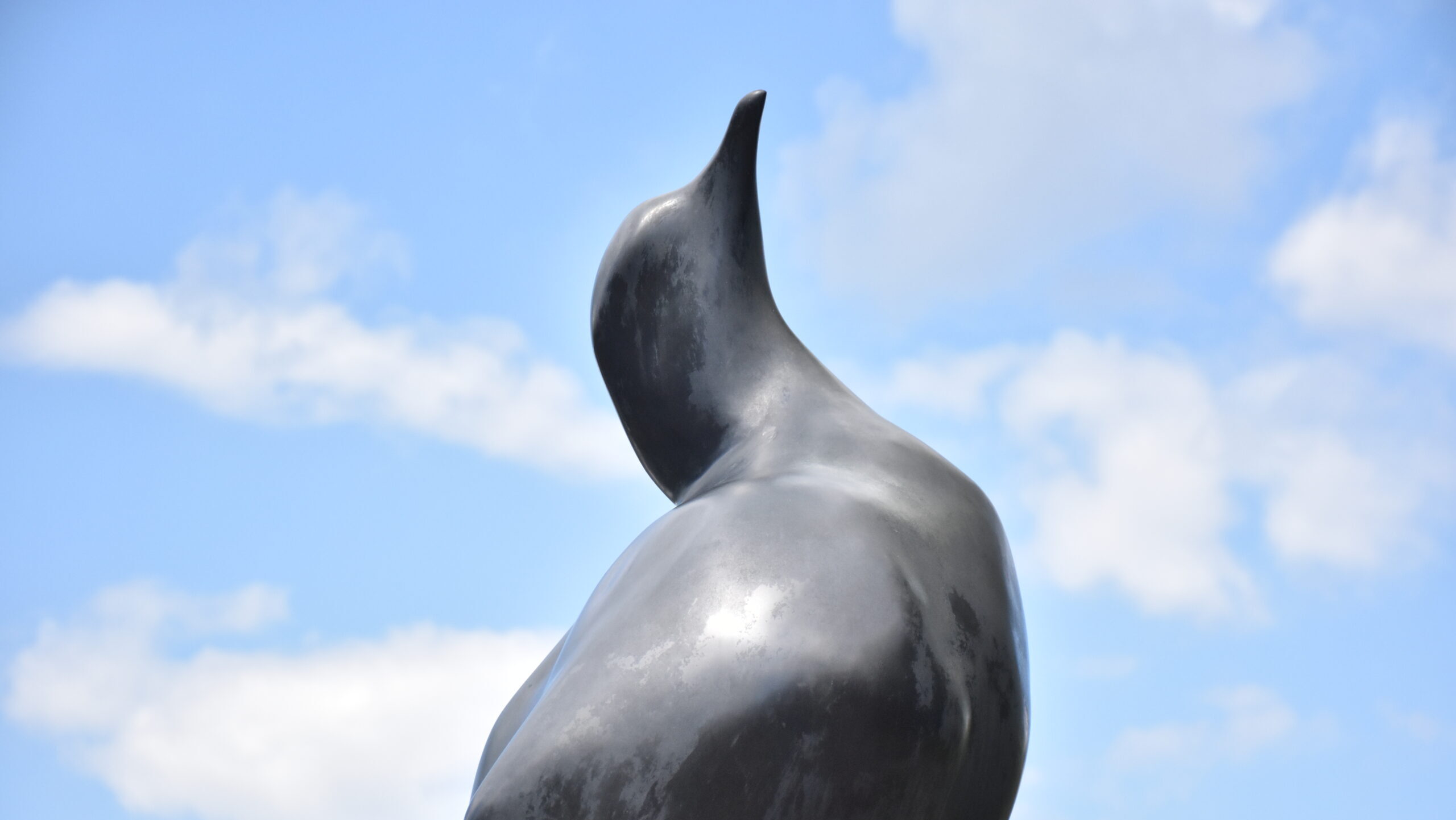 A gray metallic statue of a bird stands tall with a blue sky and white clouds in the background.