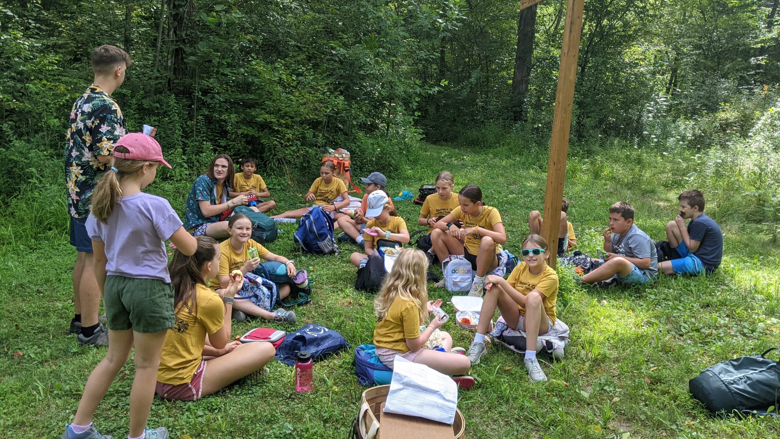 A group of campers in yellow shirts sit in a group in the grass with trees in the background