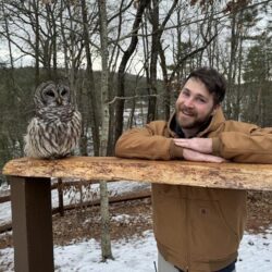 Someone in a brown jacket smiles at the camera while a barred owl perches next to them