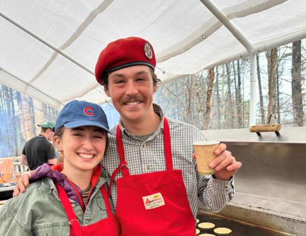 Sean wearing a red apron and red hat, posing with a co-worker in front of a griddle with pancakes.