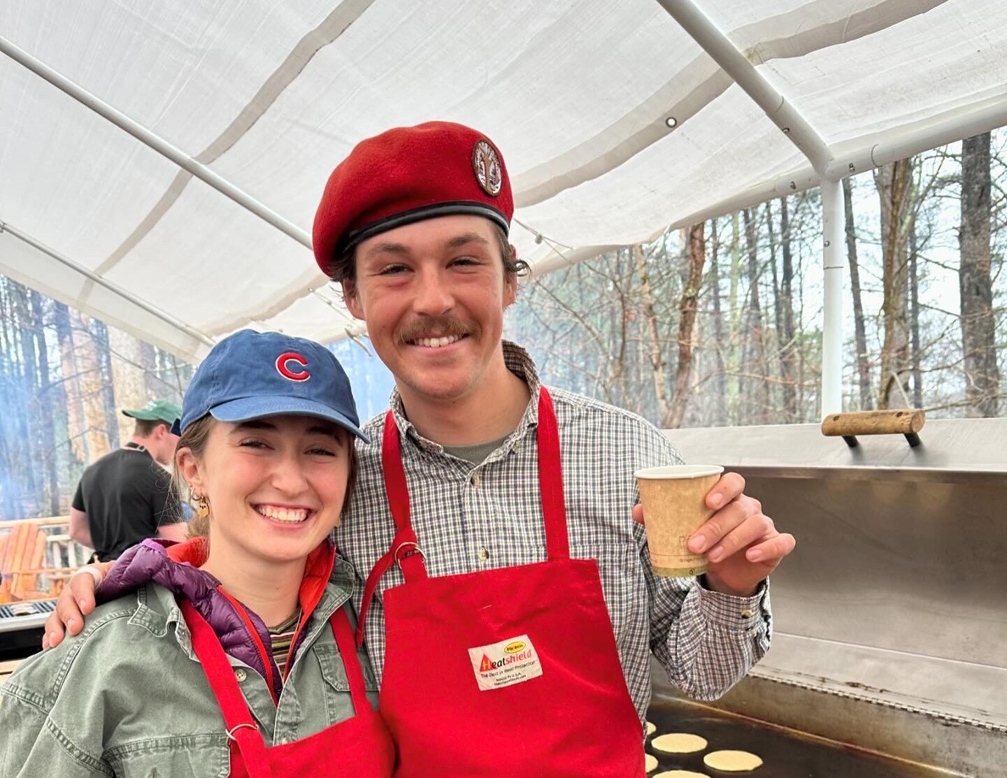 Sean wearing a red apron and red hat, posing with a co-worker in front of a griddle with pancakes.