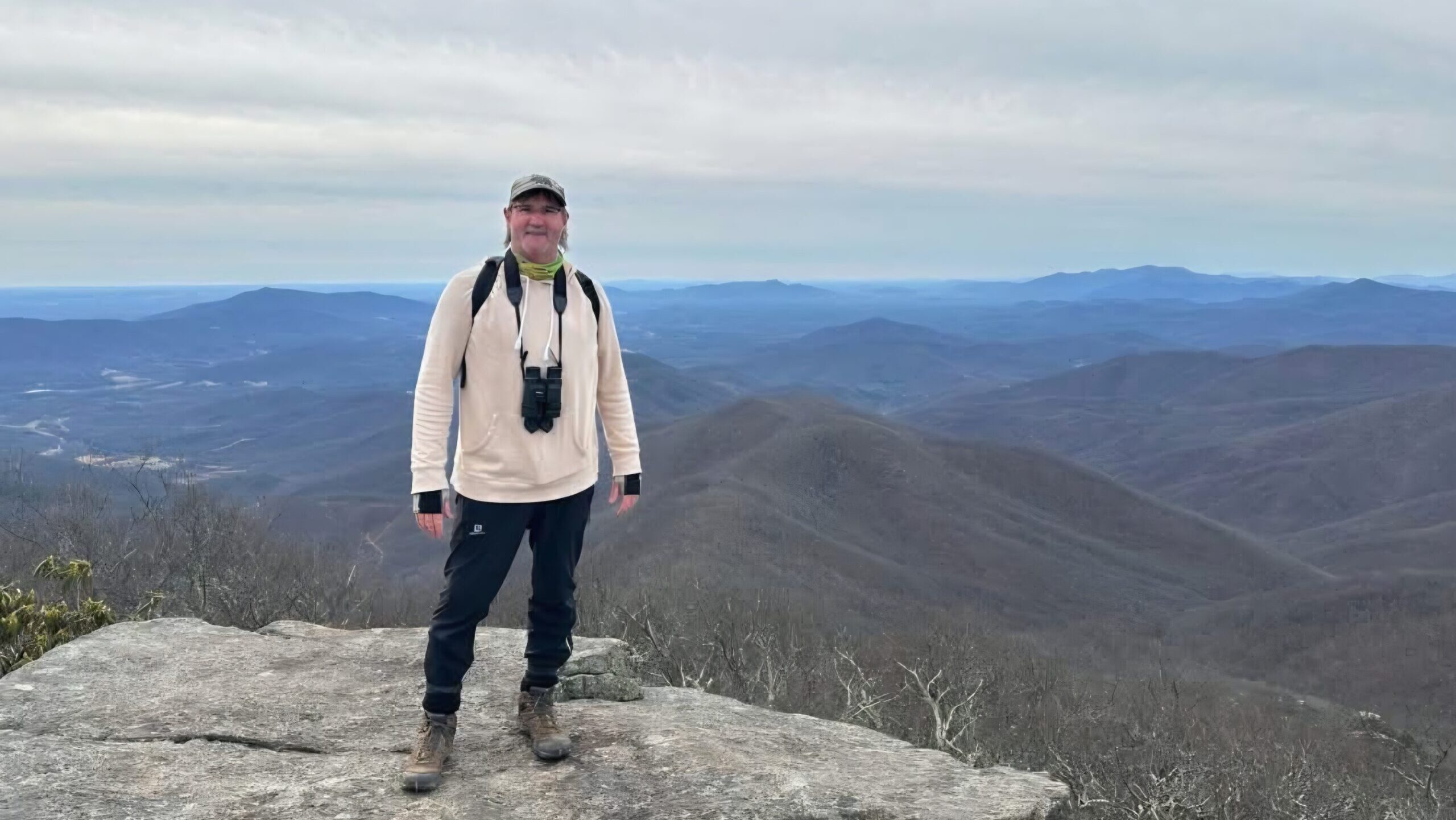 Rich Wood, wearing binoculars around his neck, stands on a rocky overlook with an expansive view of the surrounding valleys.