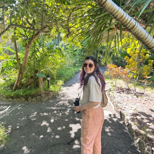 Elana posing in sunglasses with palm trees and other tropical vegetation in the background