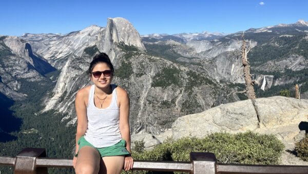 Clara sitting on a railing with a blue sky and a view of Half Dome in the background.