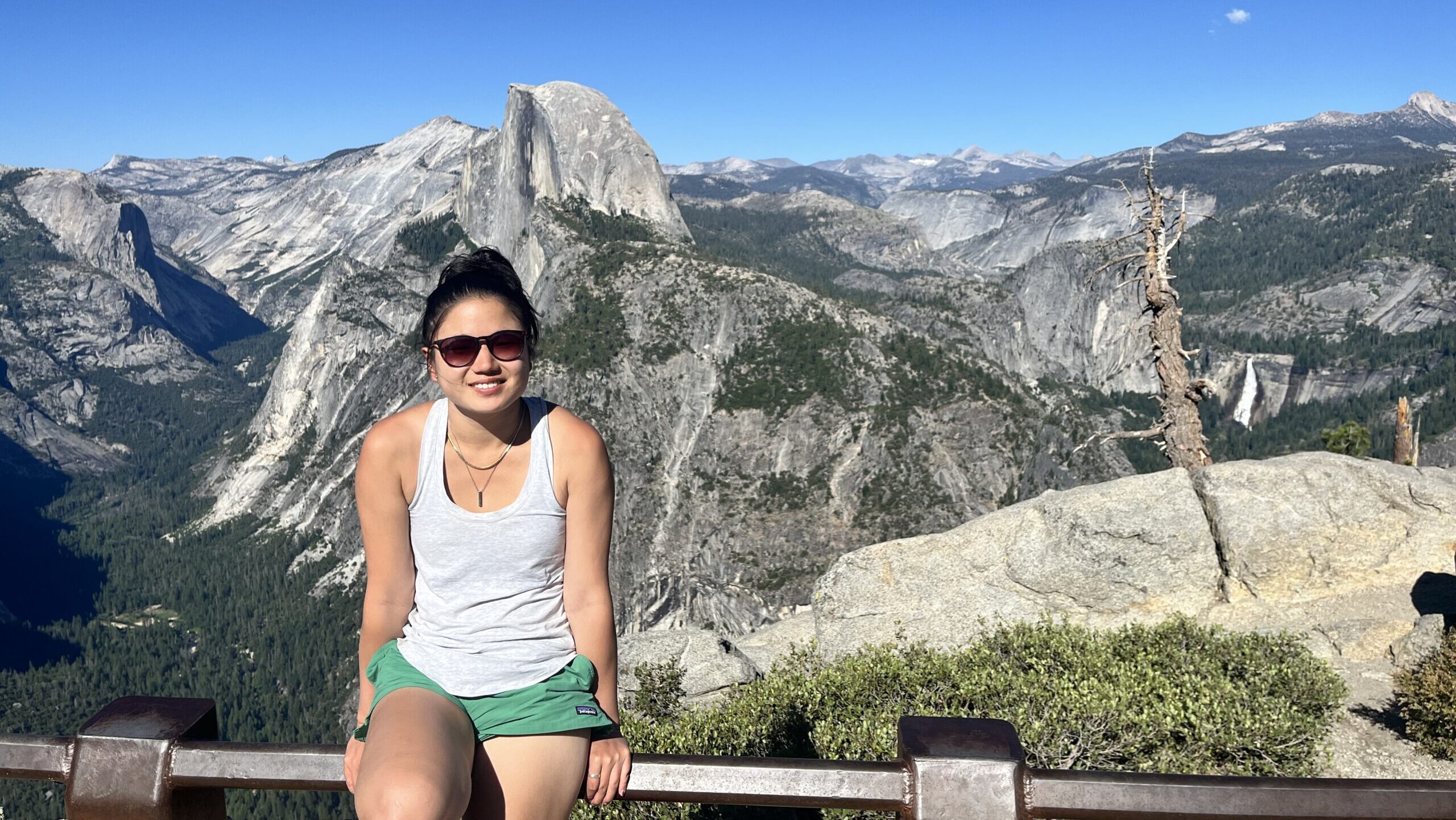 Clara sitting on a railing with a blue sky and a view of Half Dome in the background.