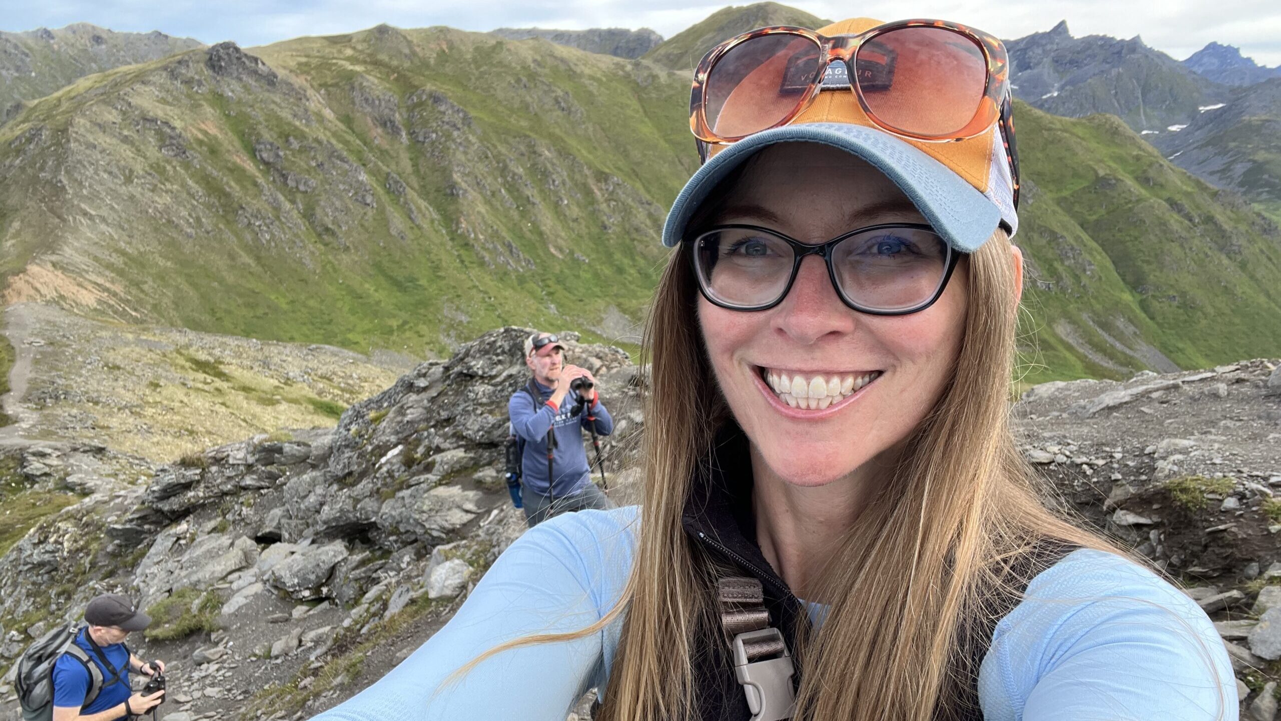 Amy, wearing sunglasses atop a baseball cap, takes a selfie on a rocky ridge with two other hikers in the background.