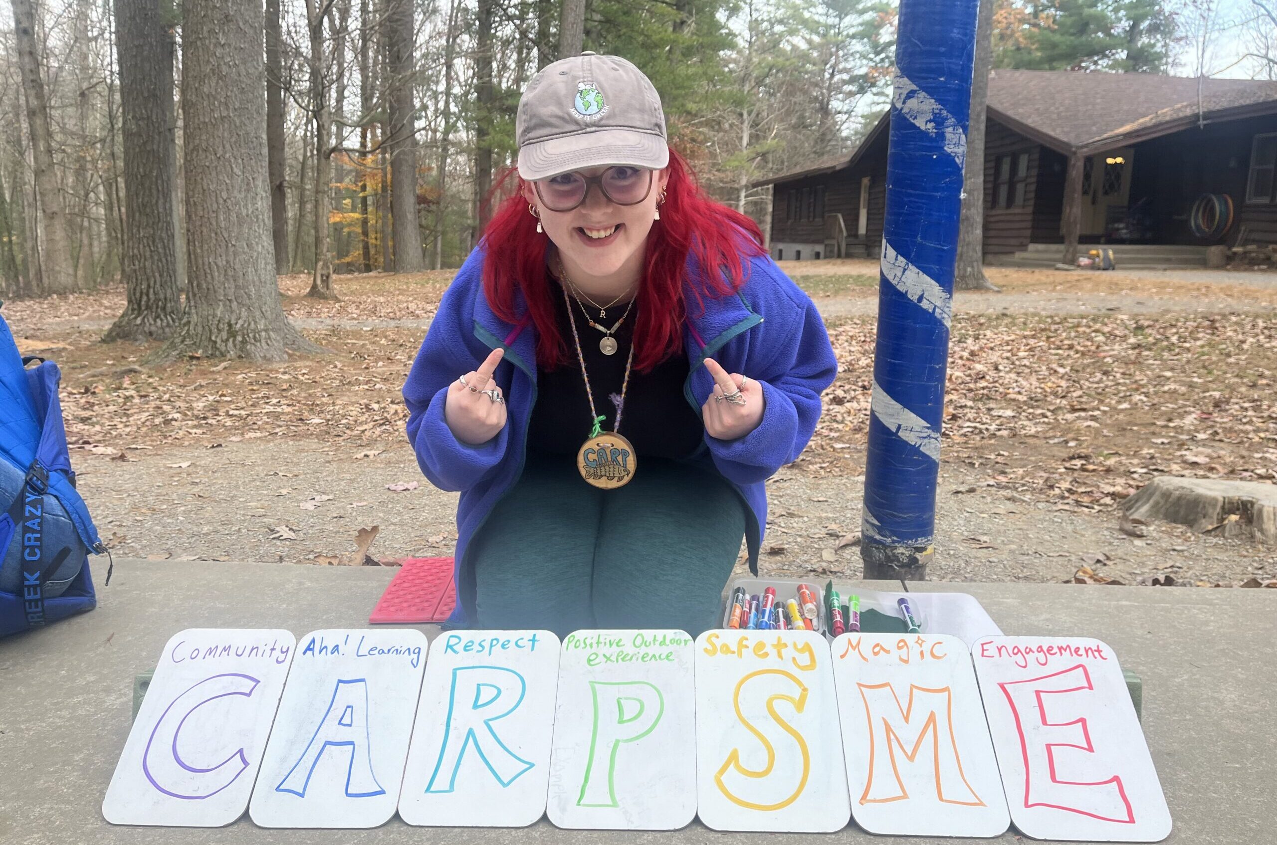 A person with dyed pink hair wearing a baseball cap kneels behind a handmade sign that has the letters CARPSME.