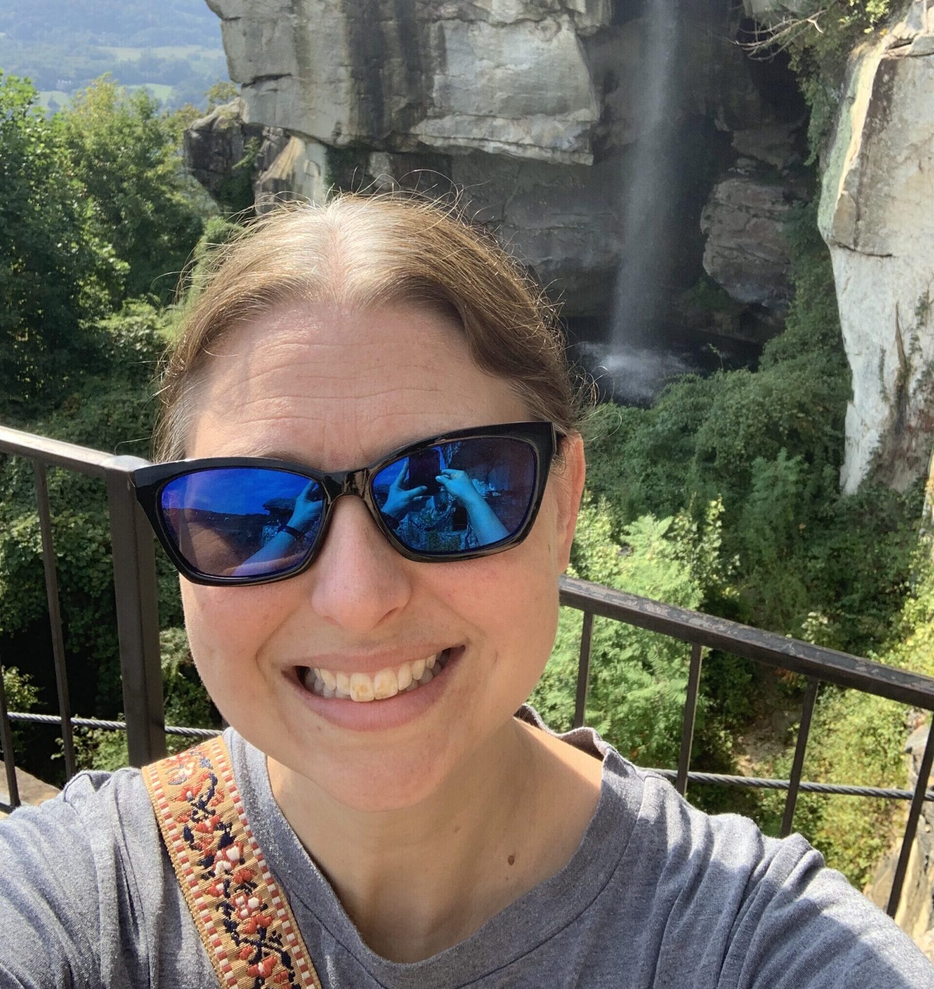 Stephanie standing in front of a rocky cliff, smiling and wearing sunglasses.