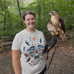 Someone in a white shirt stands in a green forest with a large bird perched on their gloved hand