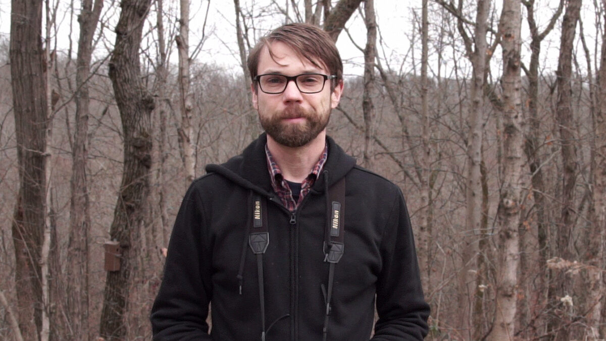 Matthew Shumar stands in the winter woods wearing binoculars around his neck.