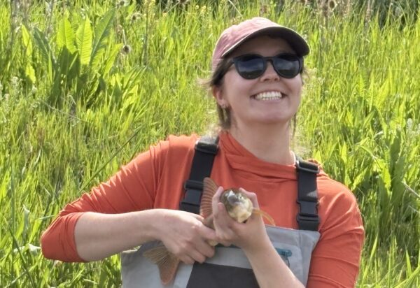 Julia wearing sunglasses, a baseball cap, and hip waders and holding a fish.