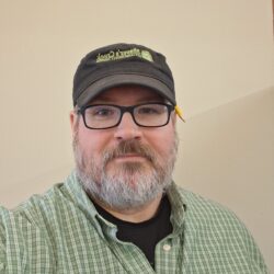 Someone wearing a green checkered shirt, glasses, and a black cap smiles at the camera in front of a white background.