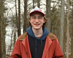 Someone wearing a red jacket and a white baseball cap smiles at the camera in front of a wooded background.
