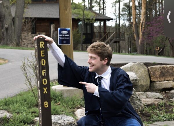 Devon, wearing a graduation gown, crouches beside a trail sign that says Red Fox.