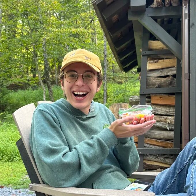 Adan relaxing in an Adirondack chair in front of a log building.