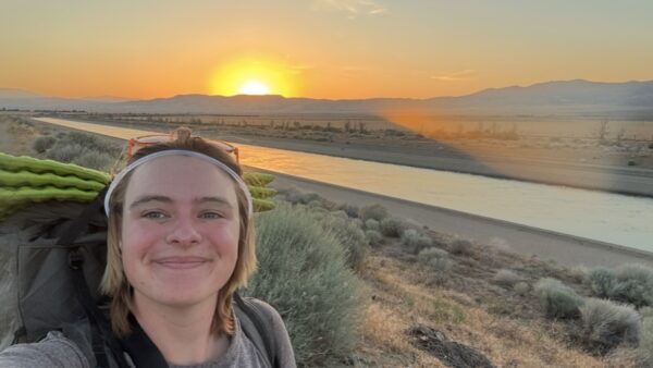 Mack smiles while taking a selfie in the sunset, wearing a backpack.