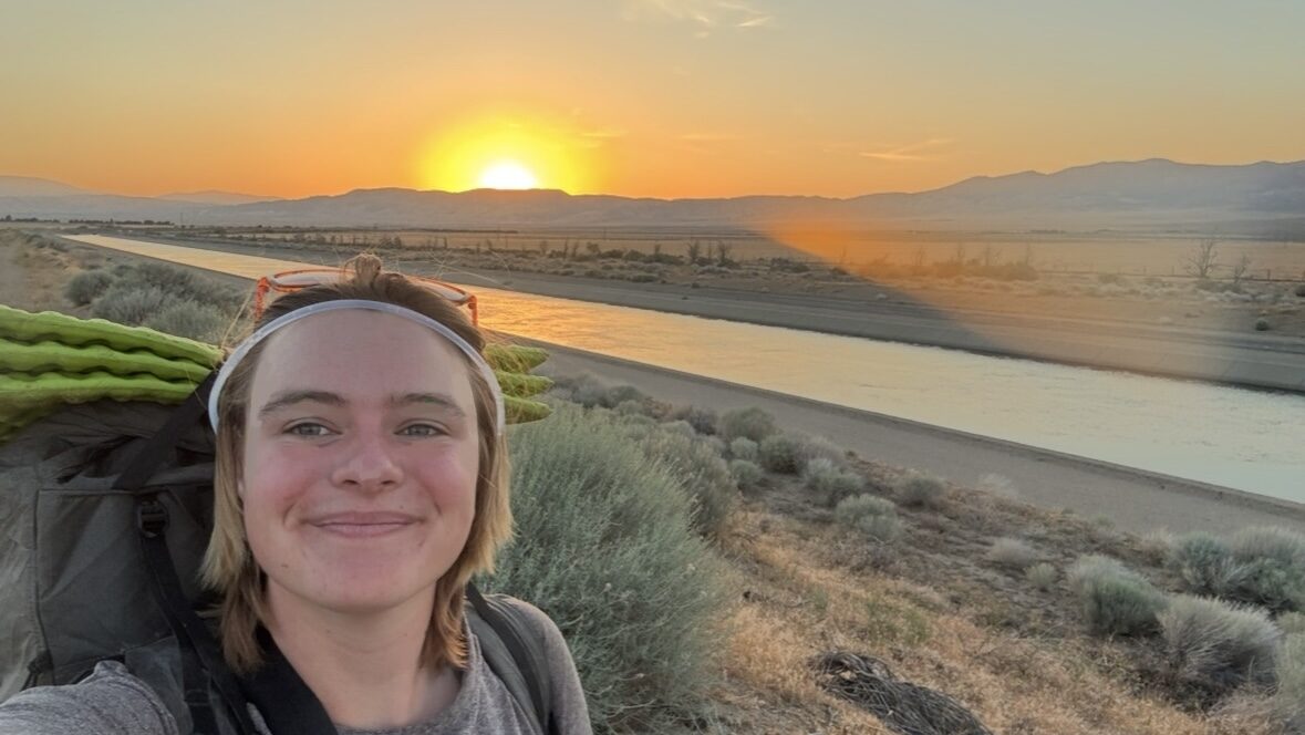Mack smiles while taking a selfie in the sunset, wearing a backpack.