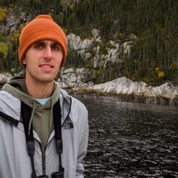 Tom Filip wearing an orange beanie and standing beside a calm, dark body of water