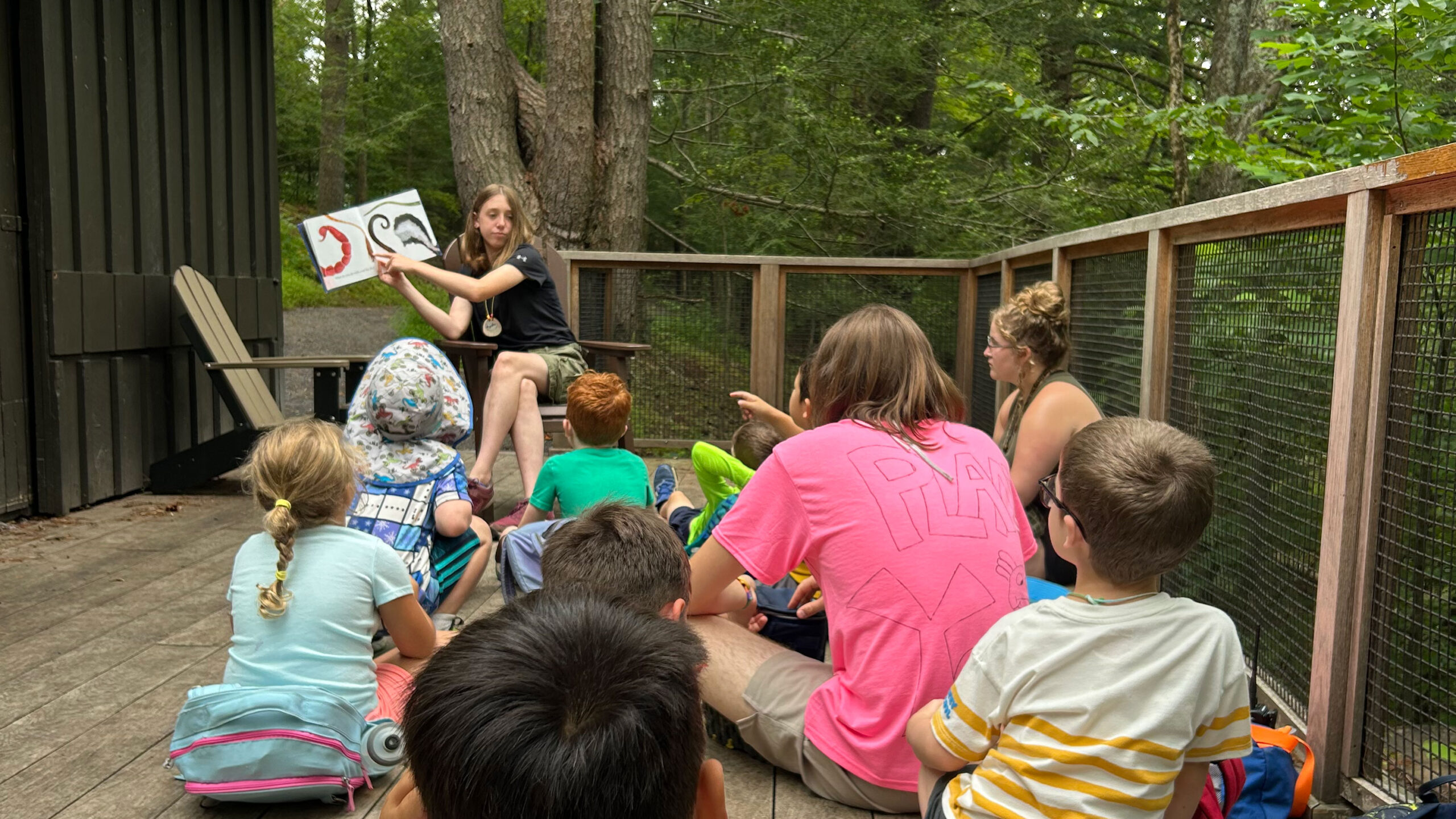 A group of children sit on a wooden deck and listen as a teen reads a story to them.
