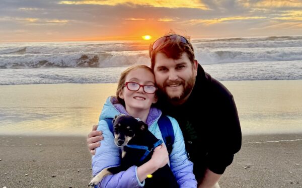 Mark and his daughter and their dog on a beach with the sun setting behind them.