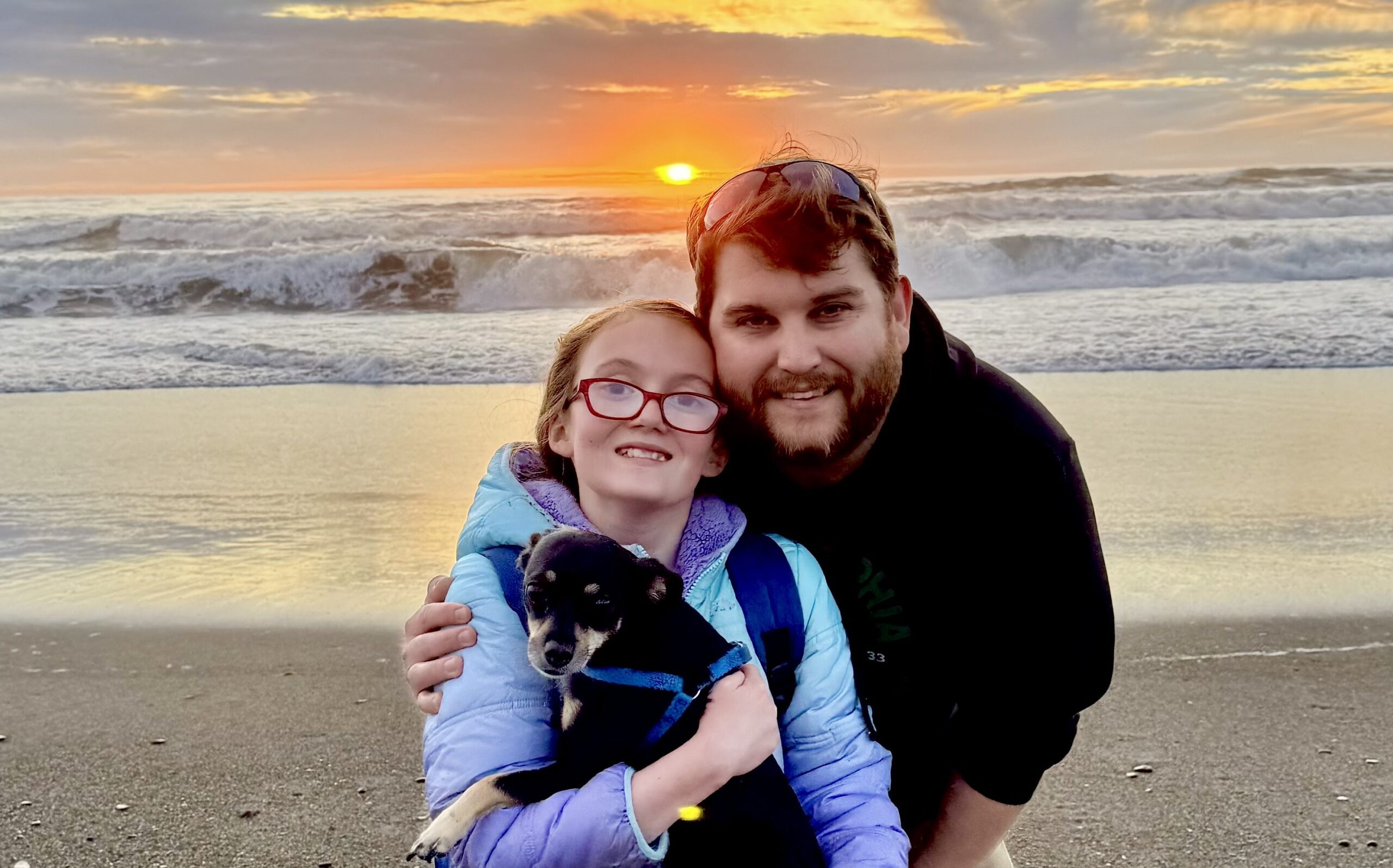 Mark and his daughter and their dog on a beach with the sun setting behind them.