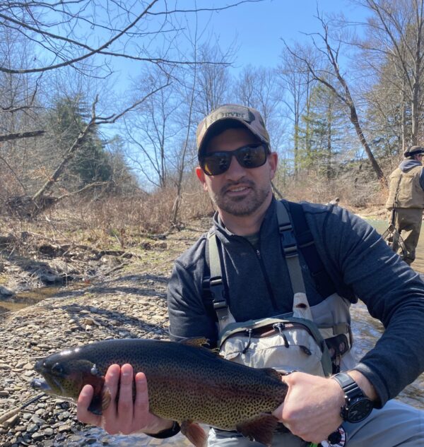 Brock wearing chest waders and kneeling in a stream while holding a large trout.