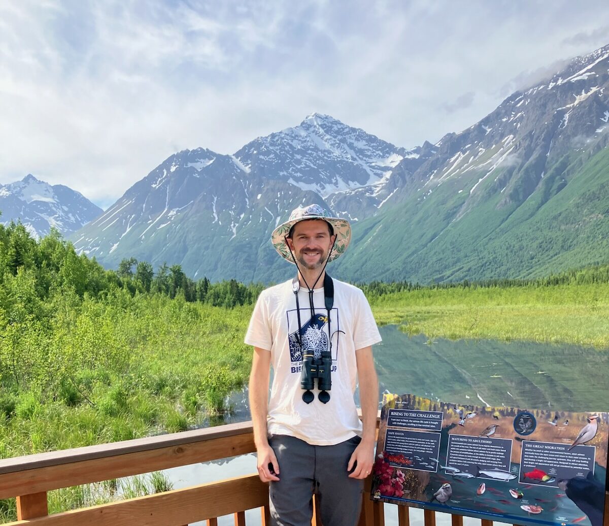 TJ standing on a deck with tall, snow-capped mountains in the distance behind him. He is wearing binoculars around his neck.